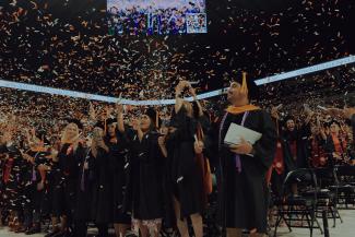 Smiling students celebrating at graduation ceremony in caps and gowns throwing confetti in the air.
